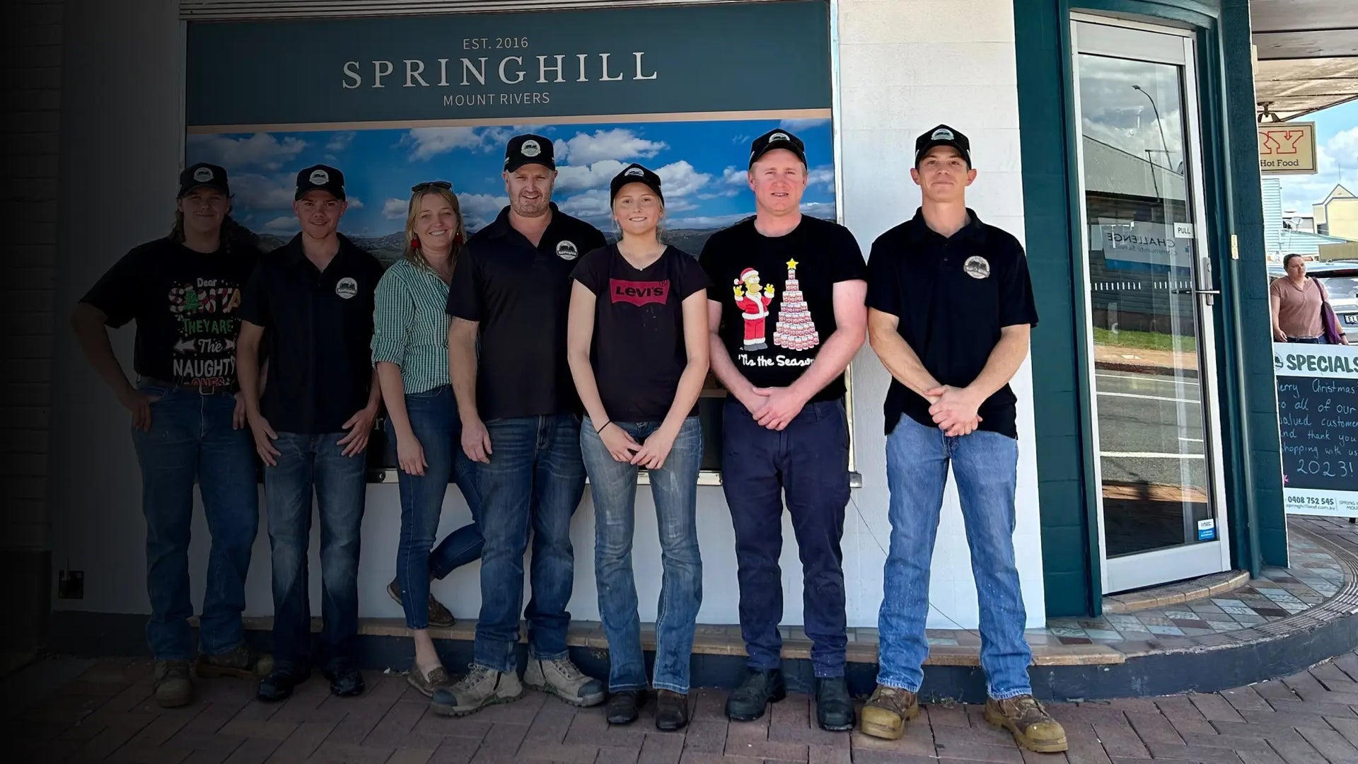 Seven men wearing matching black t-shirts and baseball caps stand in front of a building entrance with a sign reading 'Jettison Springhill Mount Rivers'.